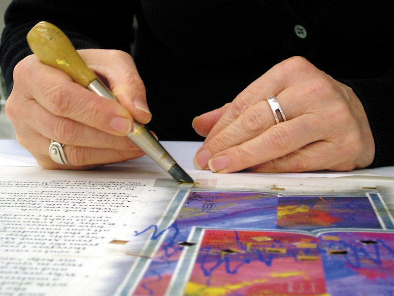 Sally Mae Joseph applies gesso over a frisket stencil in preparation for the gilding of the small squares and rectangles which appear throughout the book of Psalms. Once the gesso has dried and the gold has been laid and burnished, the frisket is pulled away, leaving perfectly sharp edges. This new technique gives Sally tremendous control gilding the geometric shapes.